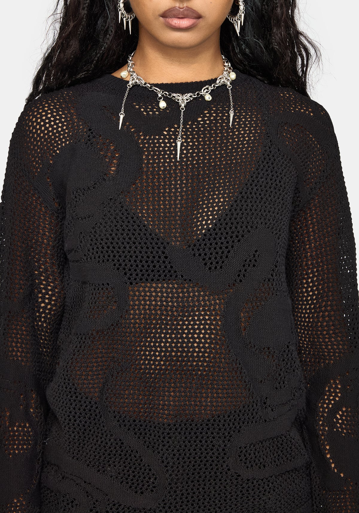 A close-up of a model wearing a black sheer crochet knit top with tonal floral lace applique overlays, styled with silver statement jewelry against a light gray background.