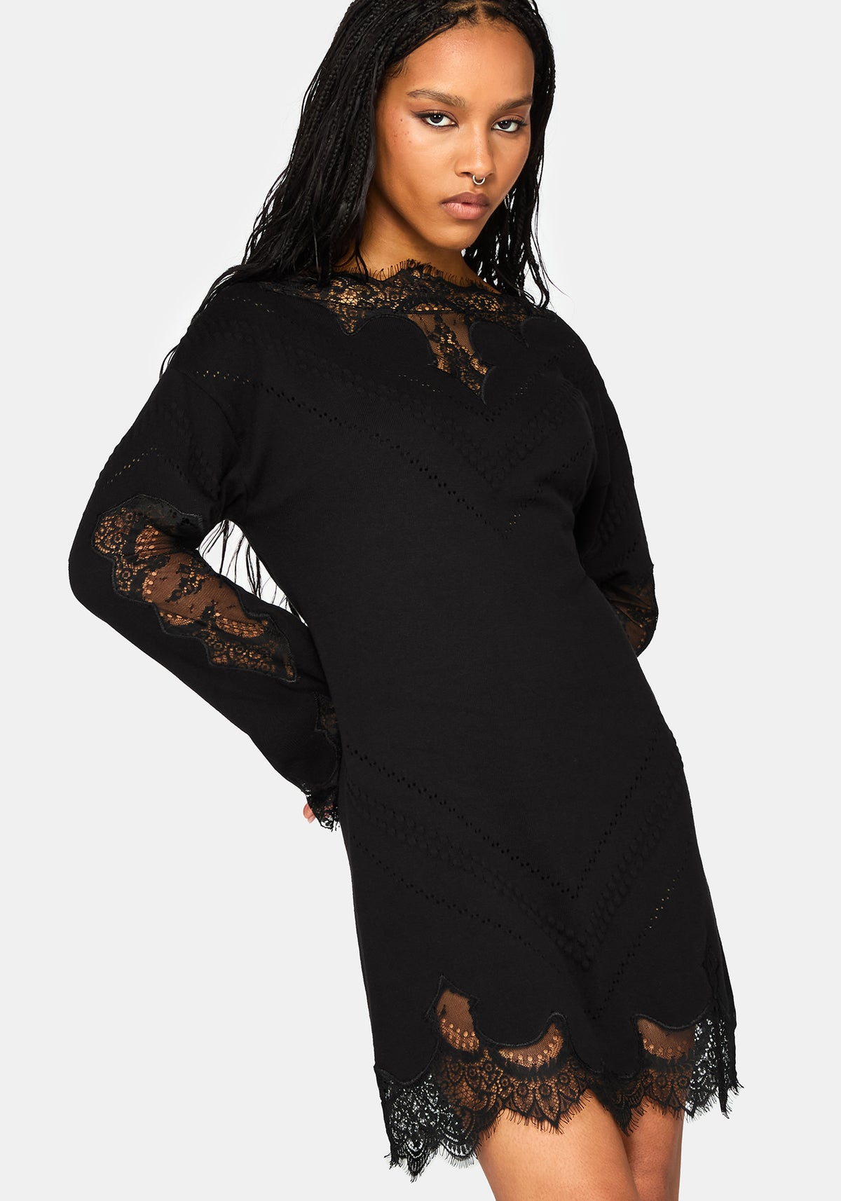 A woman modeling a black long-sleeve mini dress with intricate lace detailing on the neckline, sleeves, and hem, set against a plain light gray background.