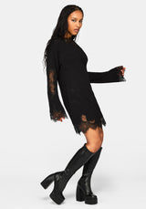 A woman modeling a black long-sleeve mini dress with sheer lace sleeves and hem, paired with black platform knee-high boots against a light gray studio background.
