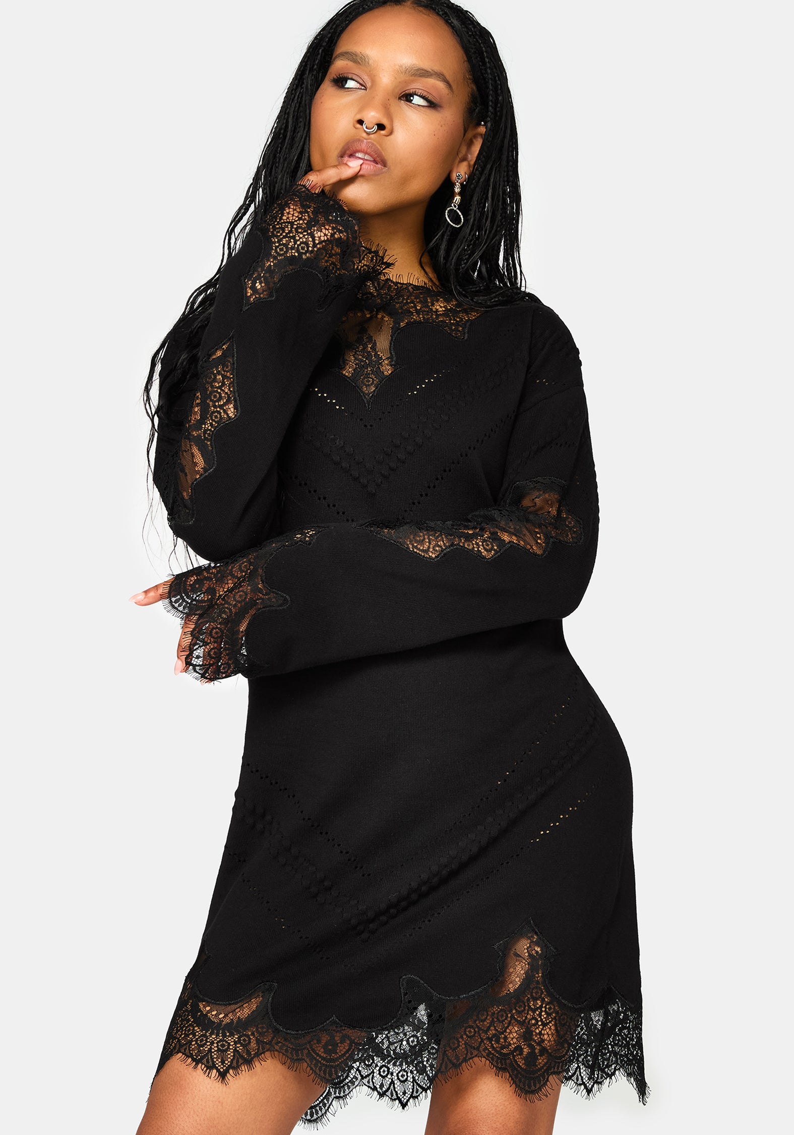 A woman modeling a black long-sleeve mini dress with sheer lace trim on the sleeves, neckline, and hem against a light white studio background.