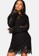 A woman modeling a black long-sleeve mini dress with sheer lace trim on the sleeves, neckline, and hem against a light white studio background.