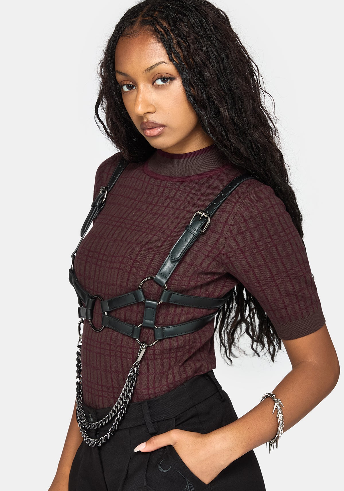 A woman wearing a dark burgundy short-sleeve top with a subtle grid pattern, styled with a black faux-leather harness vest and silver chain accents, set against a plain light gray background.