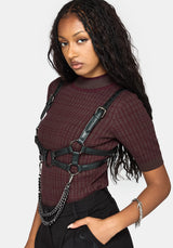 A woman wearing a dark burgundy short-sleeve top with a subtle grid pattern, styled with a black faux-leather harness vest and silver chain accents, set against a plain light gray background.