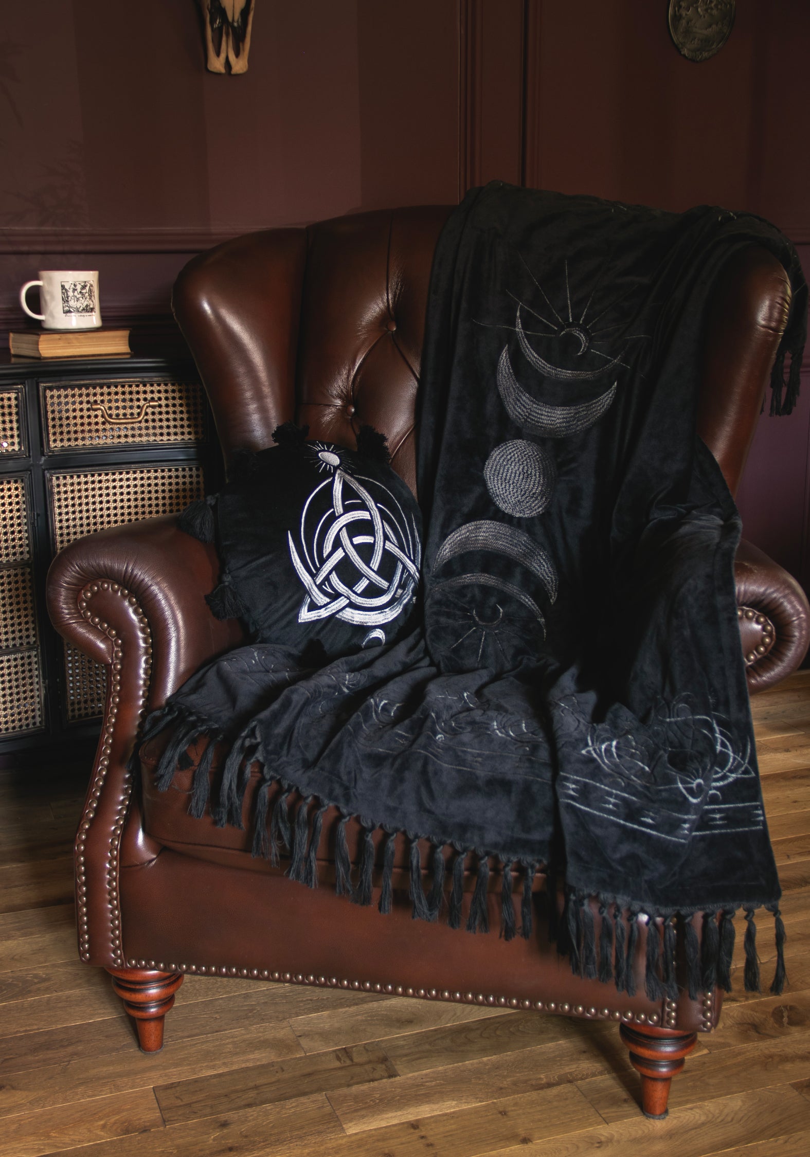 A black throw blanket and matching pillow set styled on a brown leather armchair in a cozy room. The blanket features white celestial designs including a sun, crescent moon, and full moon, along with fringe detailing.