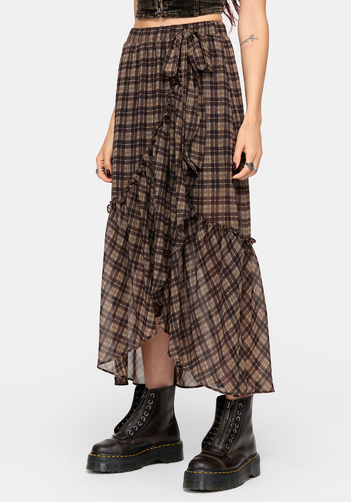 A model wearing a brown and black plaid ruffled midi skirt with a tie waist, paired with black combat boots, against a light gray studio background.