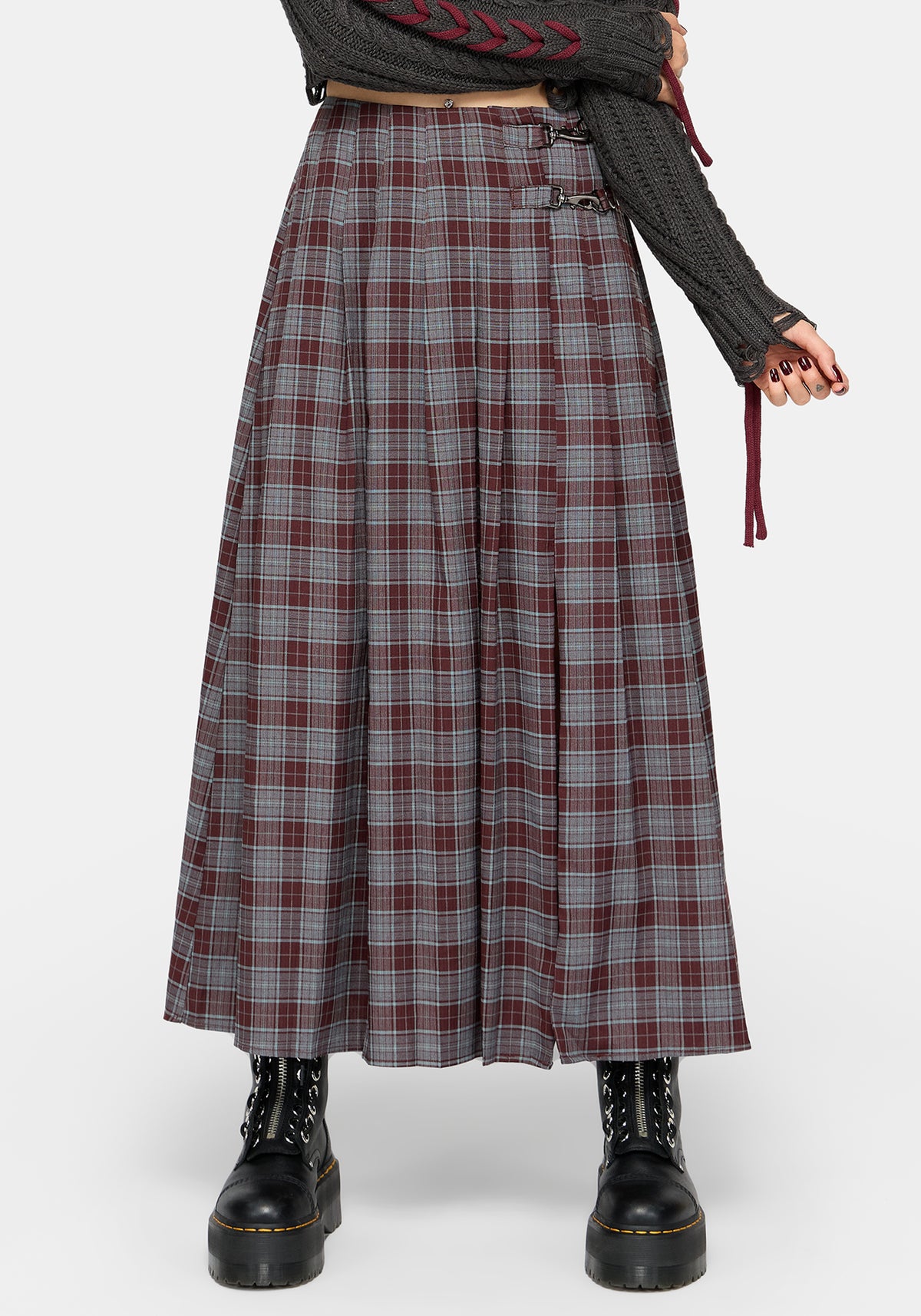 A model wearing a long, pleated plaid skirt in gray, burgundy, and white, styled with a dark gray knit sweater and black combat boots against a light gray studio background.