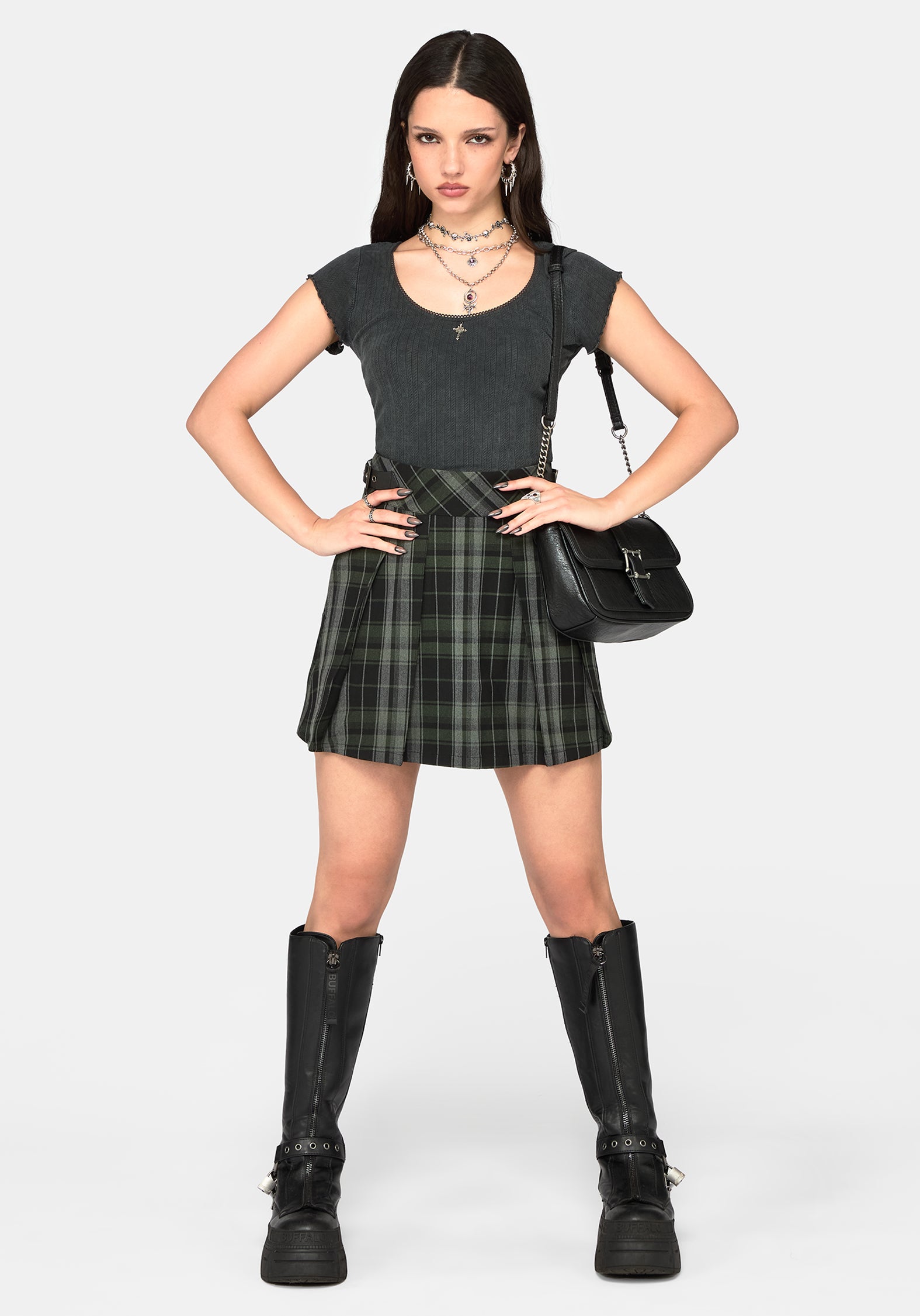 A model stands against a light gray studio background wearing a dark gray short-sleeve top, a black and green plaid pleated mini skirt, black knee-high platform boots, layered silver necklaces, and a black shoulder bag.