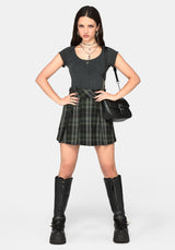 A model stands against a light gray studio background wearing a dark gray short-sleeve top, a black and green plaid pleated mini skirt, black knee-high platform boots, layered silver necklaces, and a black shoulder bag.