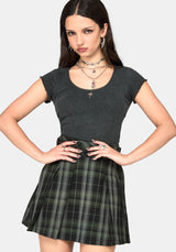 A woman posing in a dark gray ribbed short-sleeve top and a green and black plaid pleated mini skirt, accessorized with layered silver necklaces and hoop earrings, against a light gray background.