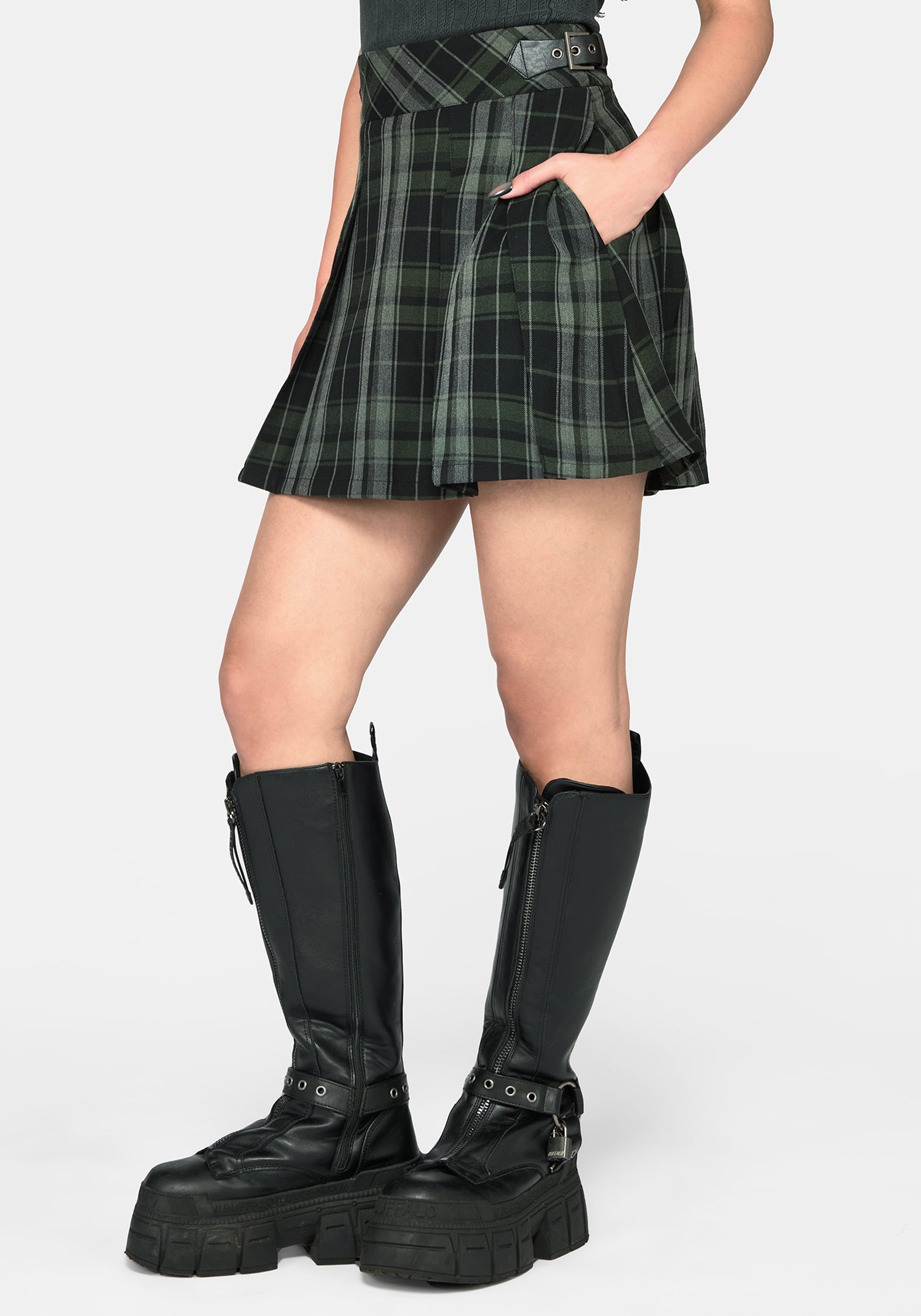A model wearing a black and green plaid pleated mini skirt with side pockets, paired with black knee-high platform boots, shown against a light gray studio background.