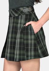 A close-up of a model wearing a dark green and black plaid pleated mini skirt with a matching belt, paired with a dark gray top against a light gray background.