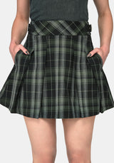 A close-up of a model wearing a pleated mini skirt in a green and black plaid pattern, with hands in the pockets, against a light gray background.