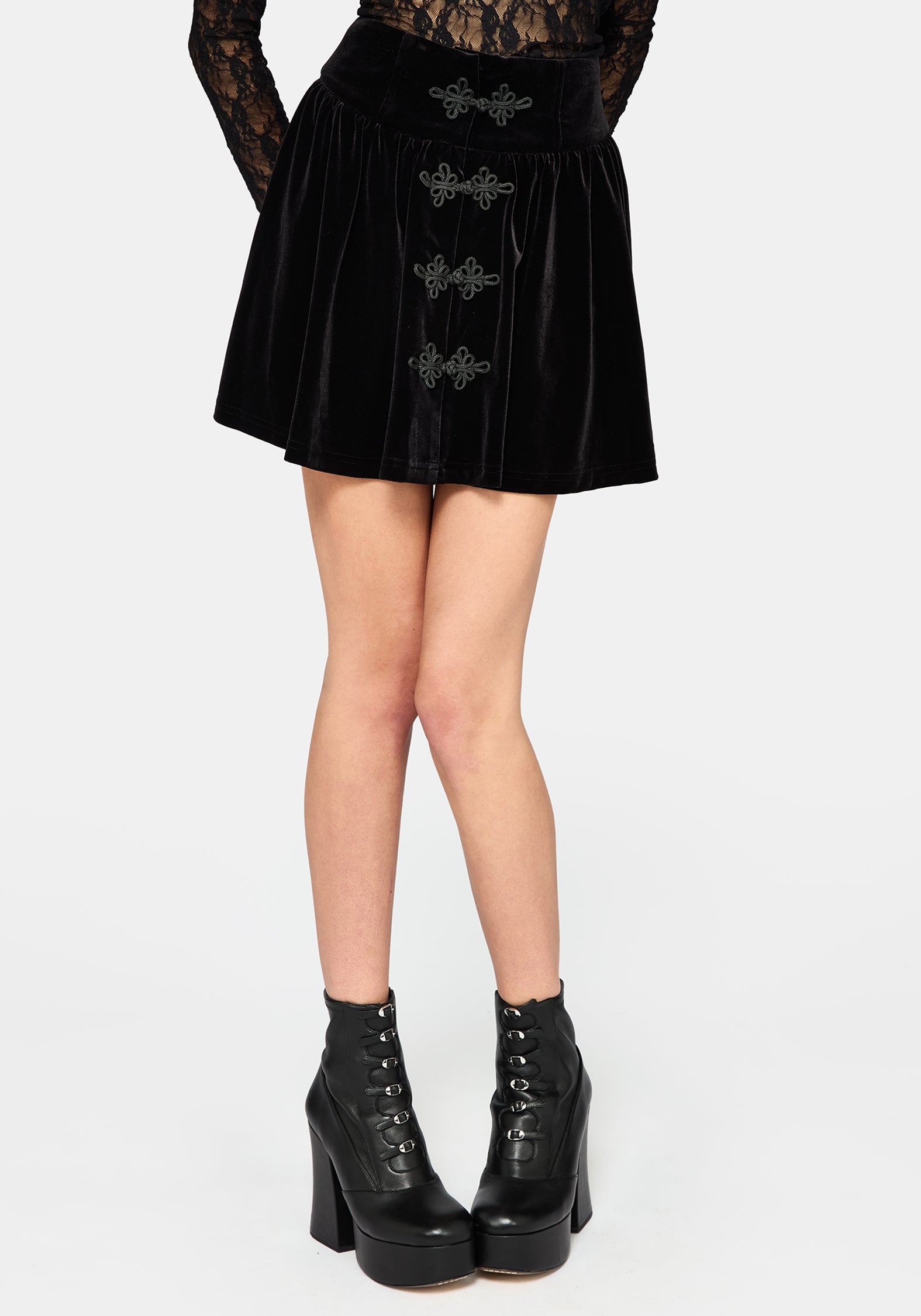 A model wearing a black velvet mini skirt with gothic silver embroidered details and lace-up ankle boots, shown from the waist down on a light gray background.