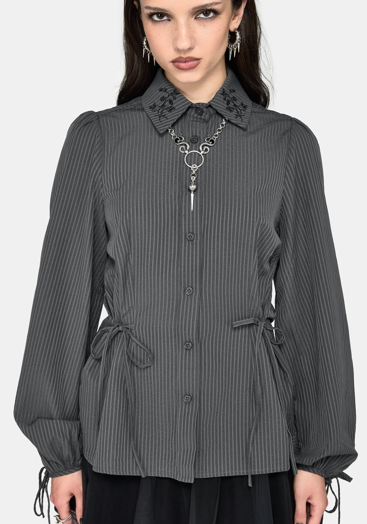 Woman wearing a dark gray pinstriped shirt with black floral embroidery on the collar, styled with a silver heart-shaped pendant necklace and silver earrings.