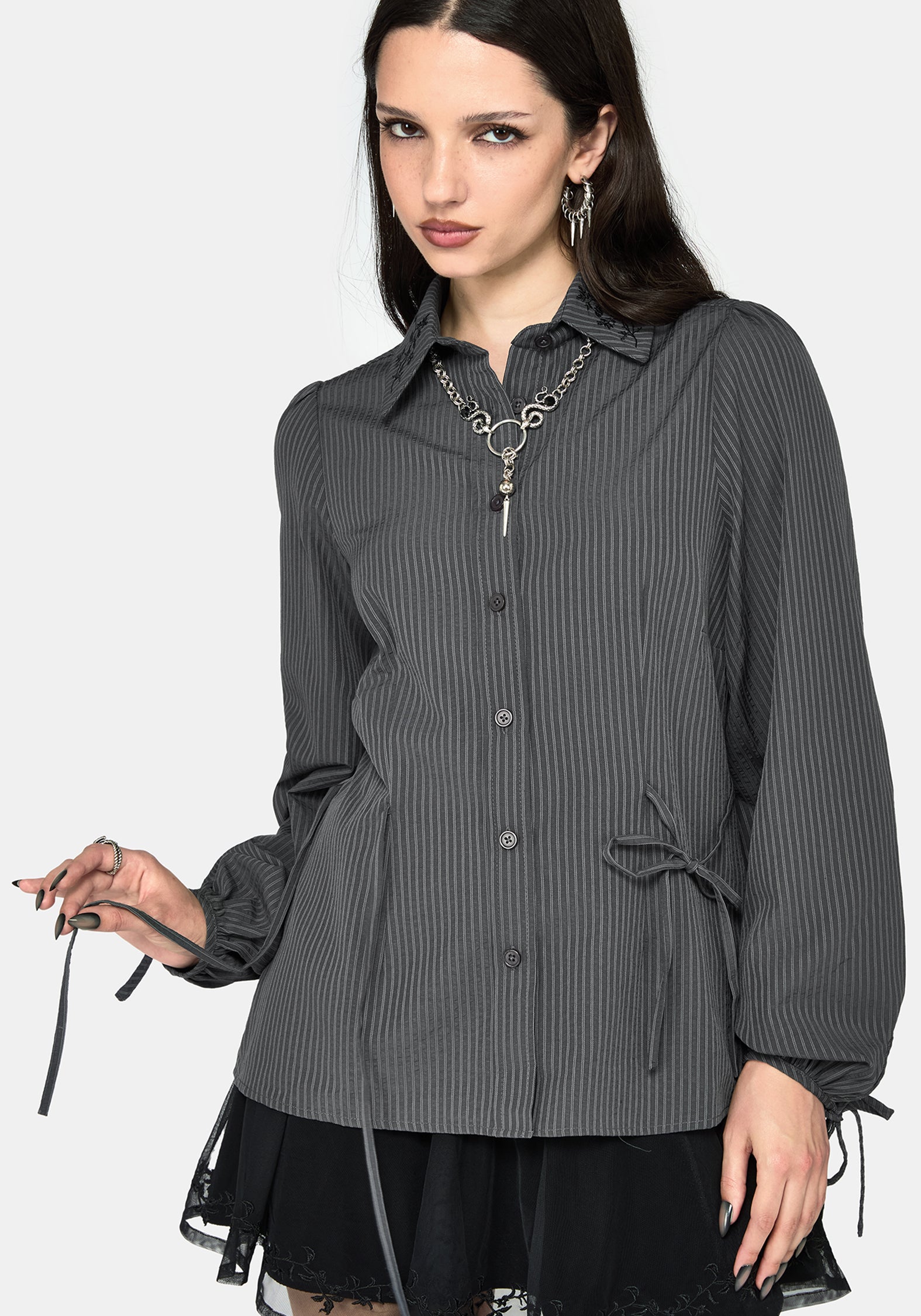 A woman modeling a dark gray and black vertically striped button-up shirt with a pointed collar, long sleeves, and side drawstrings, paired with a black lace hem dress against a light gray background.