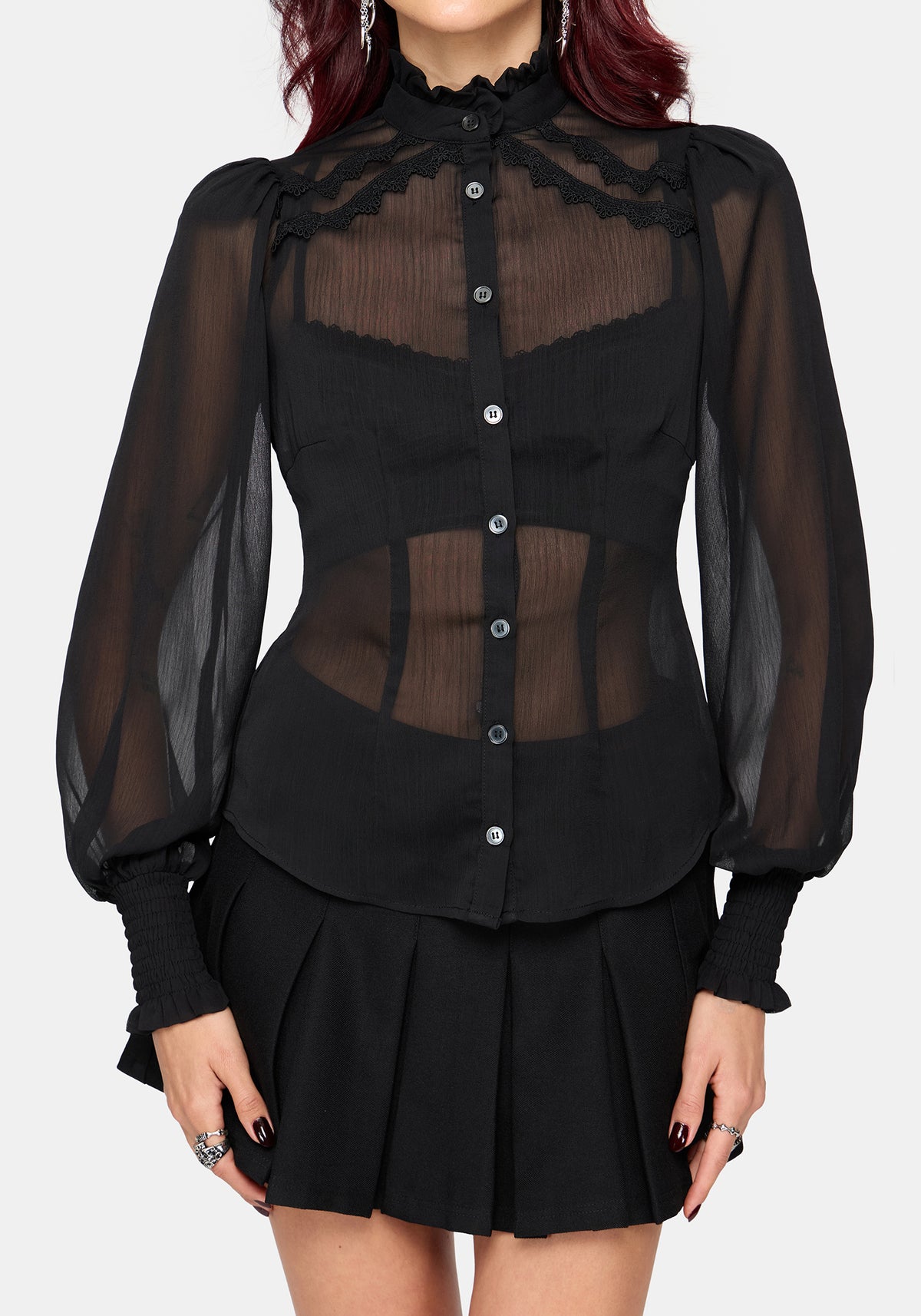 A model wearing a black sheer long-sleeve blouse with lace trim and a black pleated mini skirt, shown against a light gray studio background.