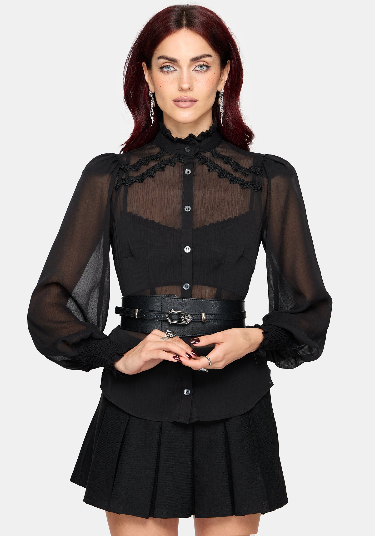 A woman modeling a black long-sleeve blouse with sheer mesh sleeves, a high ruffled collar, and a sheer yoke panel, paired with a matching pleated black skirt and a black belt with silver hardware, against a light gray background.
