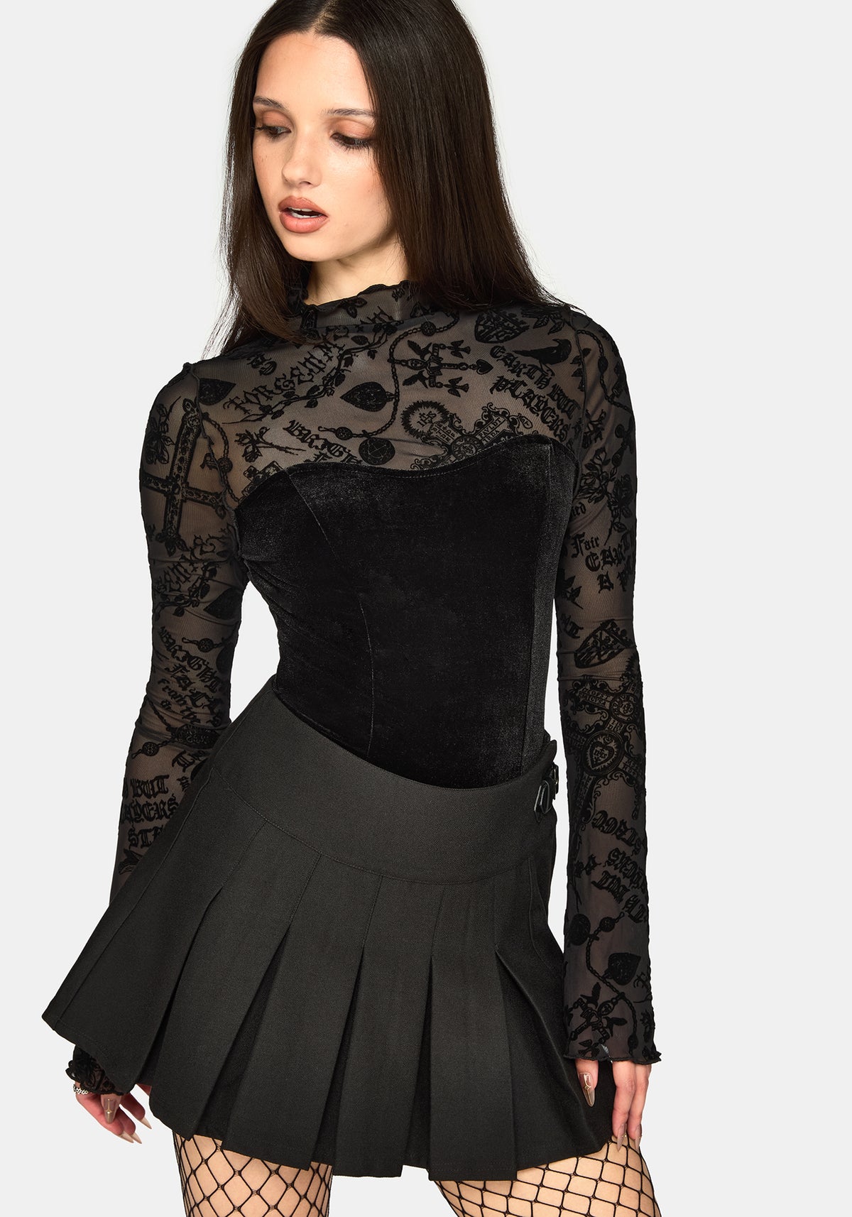 A model wearing a black long-sleeve mock-neck top with sheer floral mesh sleeves and a black velvet corset-style bodice, paired with a black pleated mini skirt and fishnet stockings, against a light gray background.
