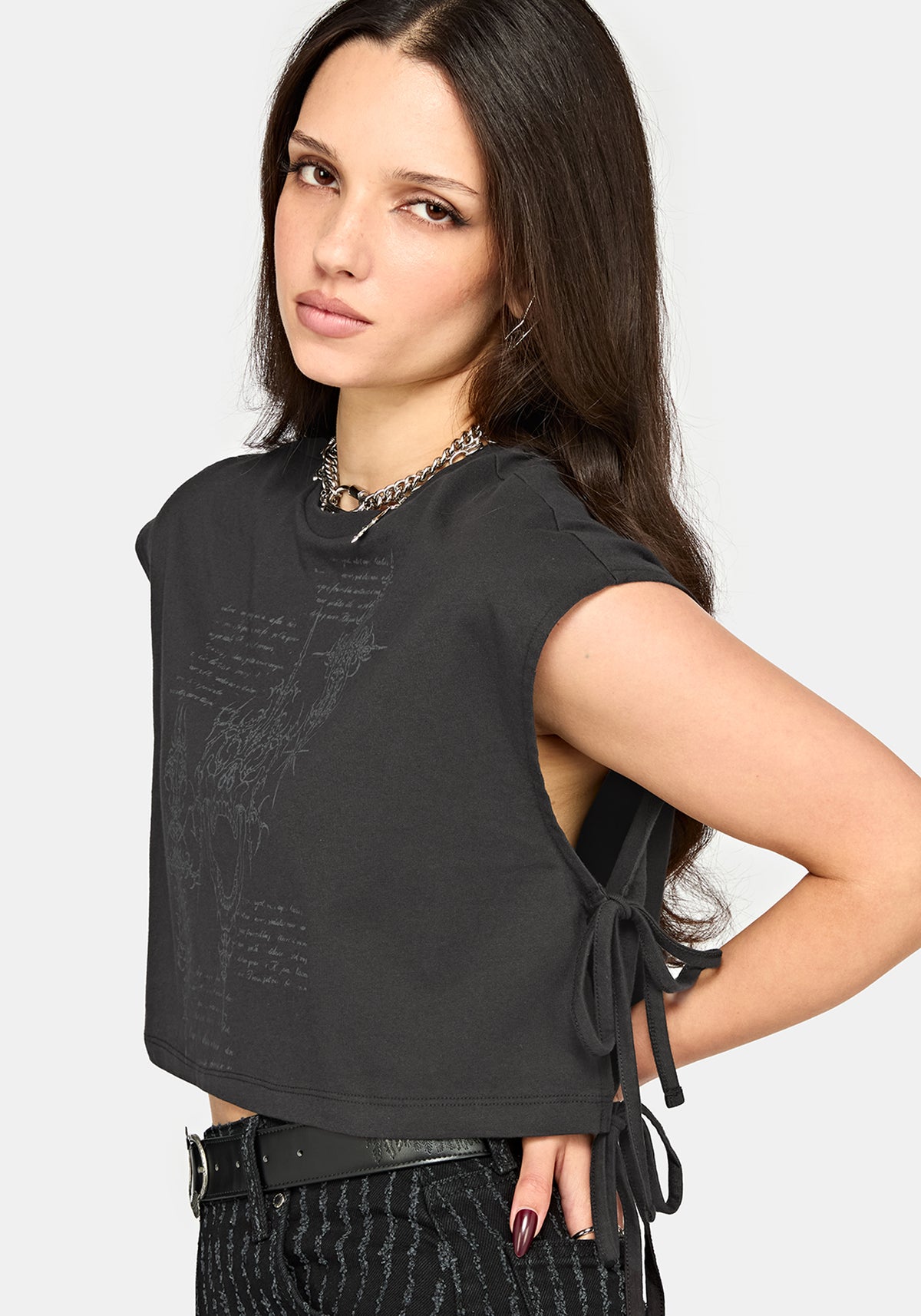 A woman modeling a black cropped sleeveless top with a subtle tonal graphic print on the front and side tie details, paired with black patterned pants against a light gray studio background.