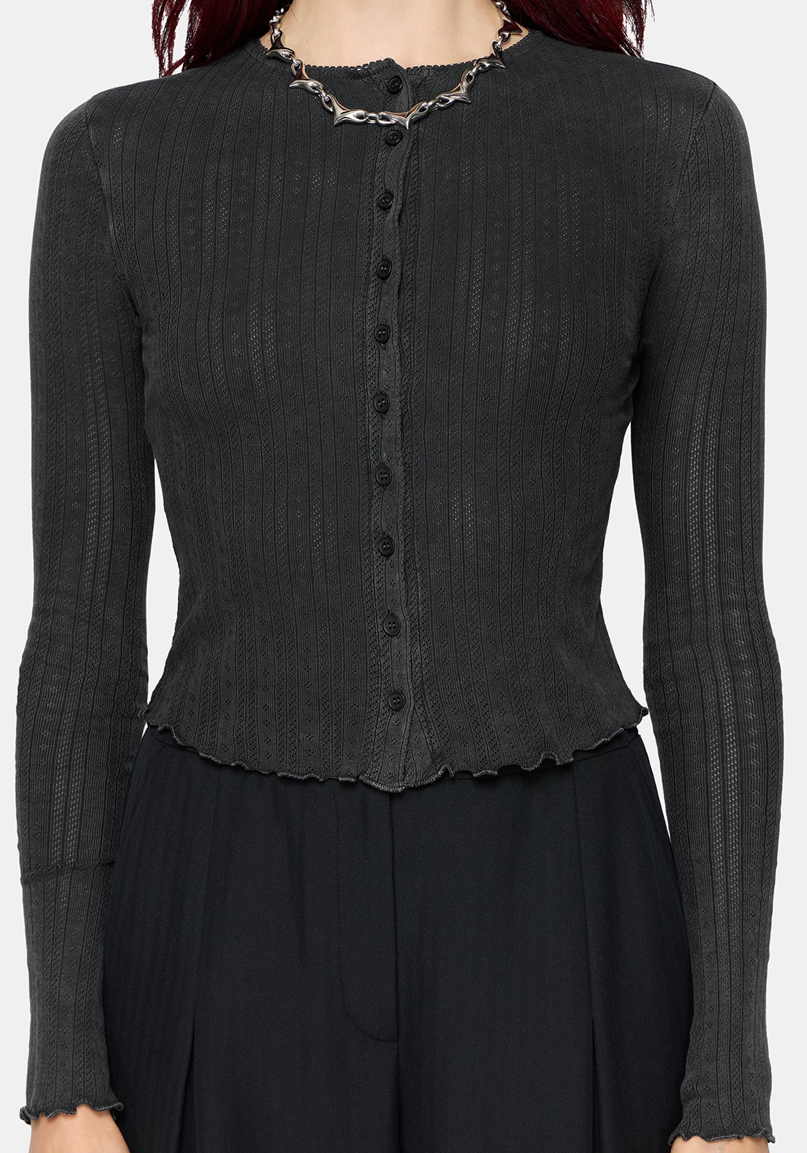 A woman wearing a dark charcoal gray ribbed long-sleeve cardigan with a button-front detail and scalloped hem, styled with a chunky silver chain necklace against a light gray background.
