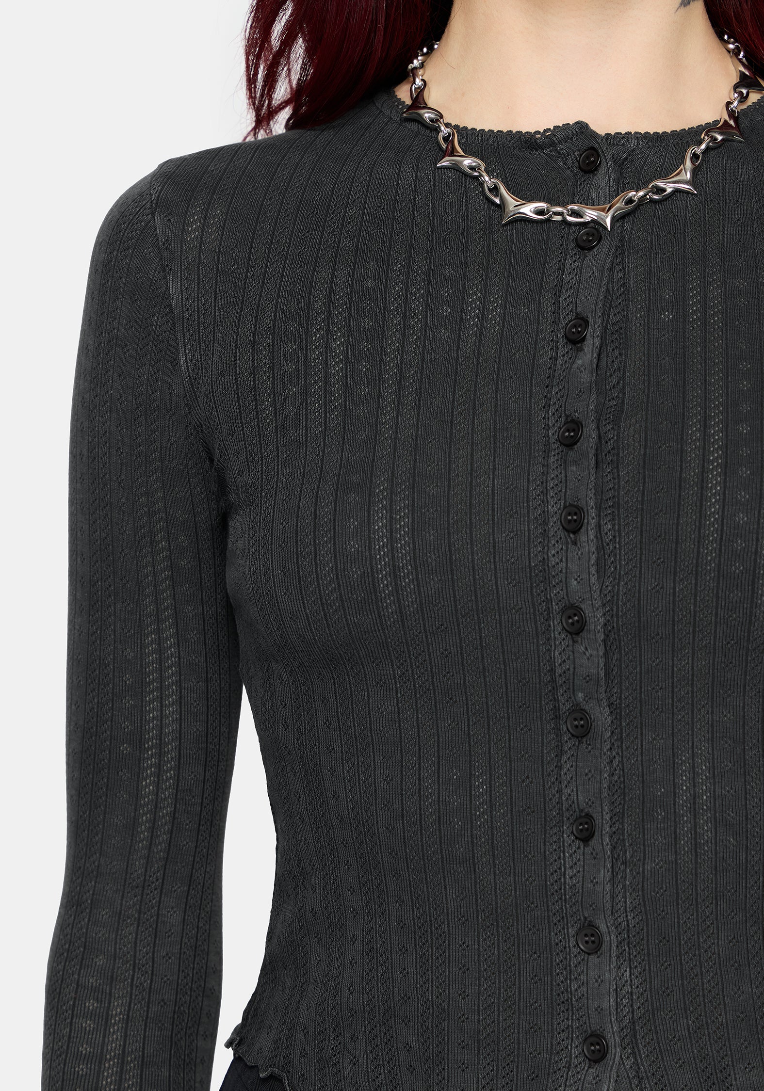 A woman wearing a dark charcoal gray ribbed long-sleeve cardigan with a button-front detail and scalloped hem, styled with a chunky silver chain necklace against a light gray background.
