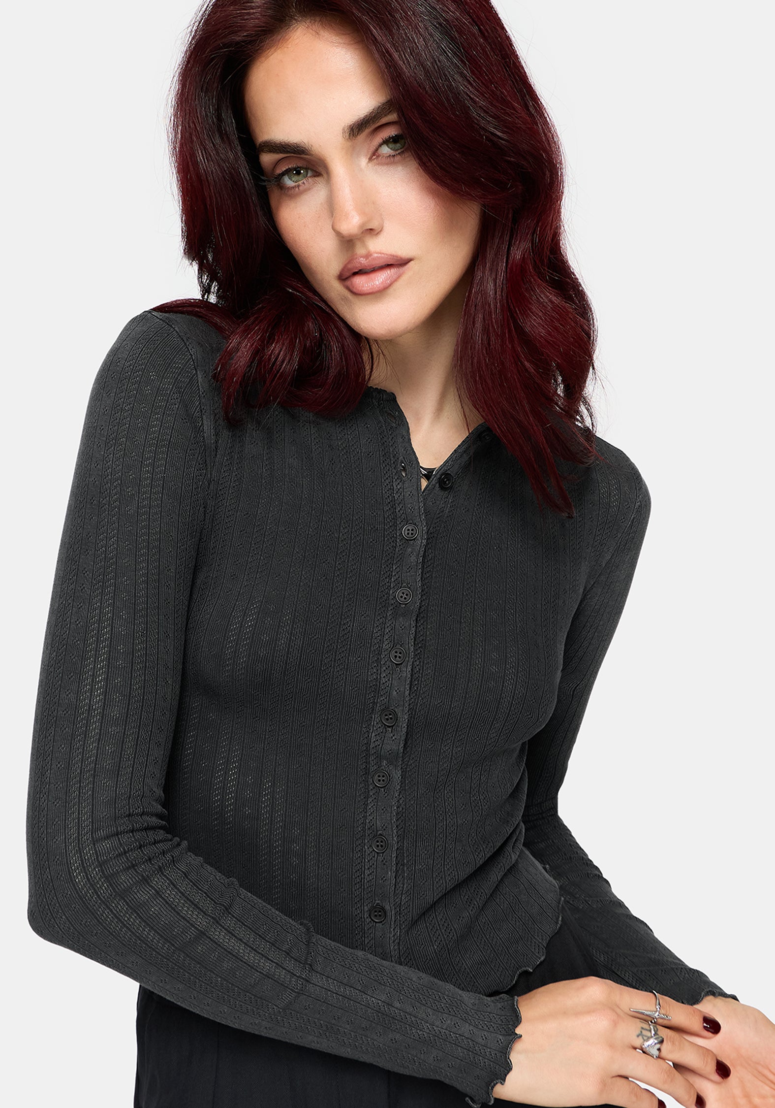 A woman with dark red hair wearing a dark charcoal gray ribbed long-sleeve button-up knit top, photographed against a light gray studio background.