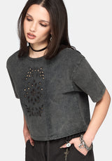 A woman wearing a dark gray washed short-sleeve crop top with a decorative black embroidered panel on the front, styled with black pants and accessories against a light gray background.
