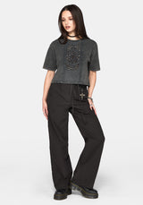 A woman wearing a dark gray washed short-sleeve crop top with a vertical panel of cut out details and embroidery, paired with black pants and a black choker against a light grey background.