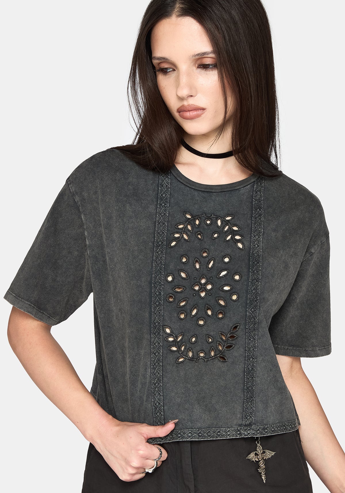 A woman wearing a dark gray washed short-sleeve crop top with a vertical panel of cut out details and embroidery, paired with black pants and a black choker against a light grey background.