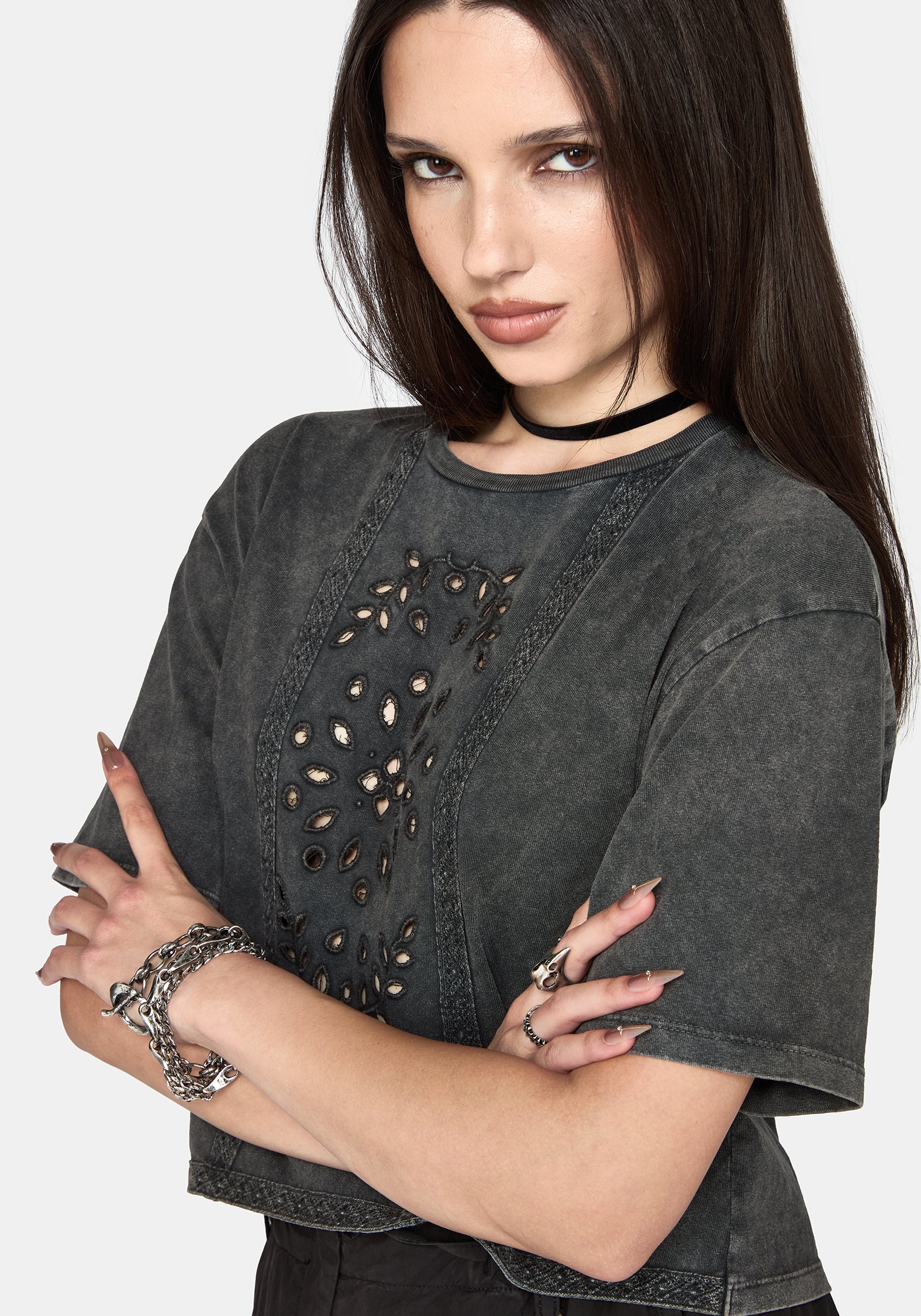 A woman wearing a dark grey washed short-sleeve crop top with cutout details on the front, styled with a black choker and silver bracelets against a light gray background.