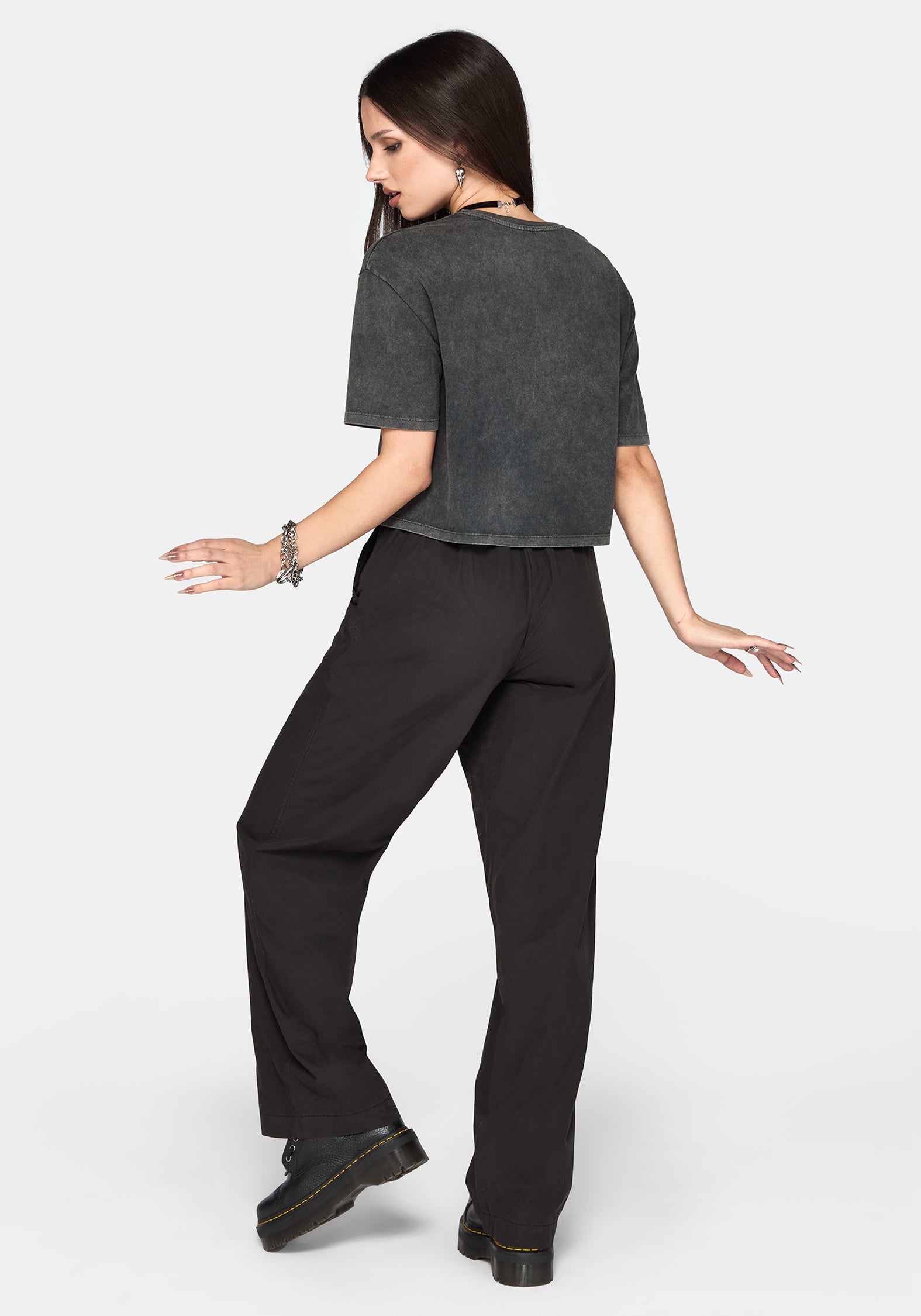 A woman posing from the back wearing a dark grey cropped t-shirt and black wide-leg trousers against a light gray background.