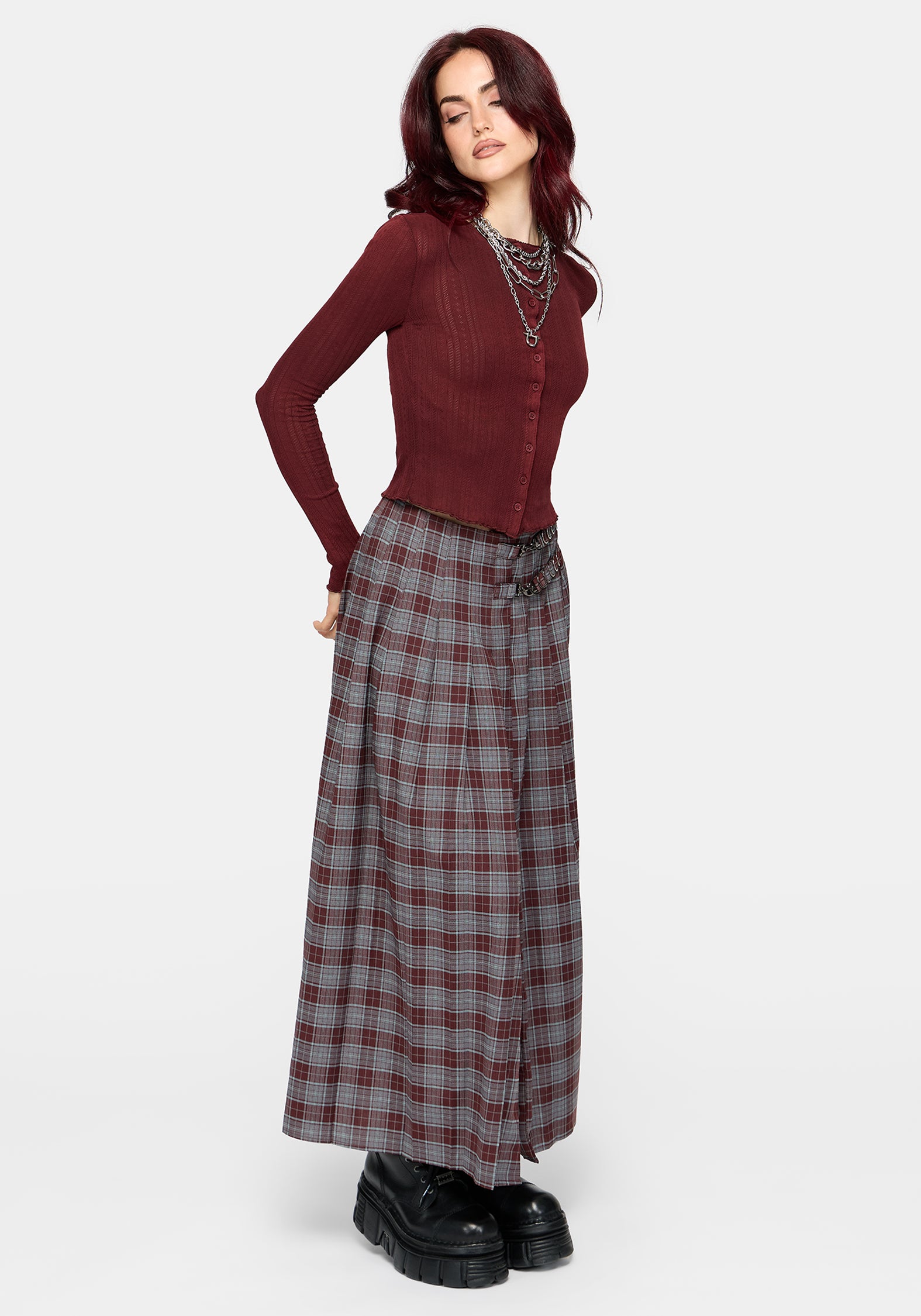 A woman wearing a ribbed long-sleeve button-front knit top in deep burgundy, paired with a gray and burgundy plaid skirt and layered silver chain necklaces, against a light gray studio background.