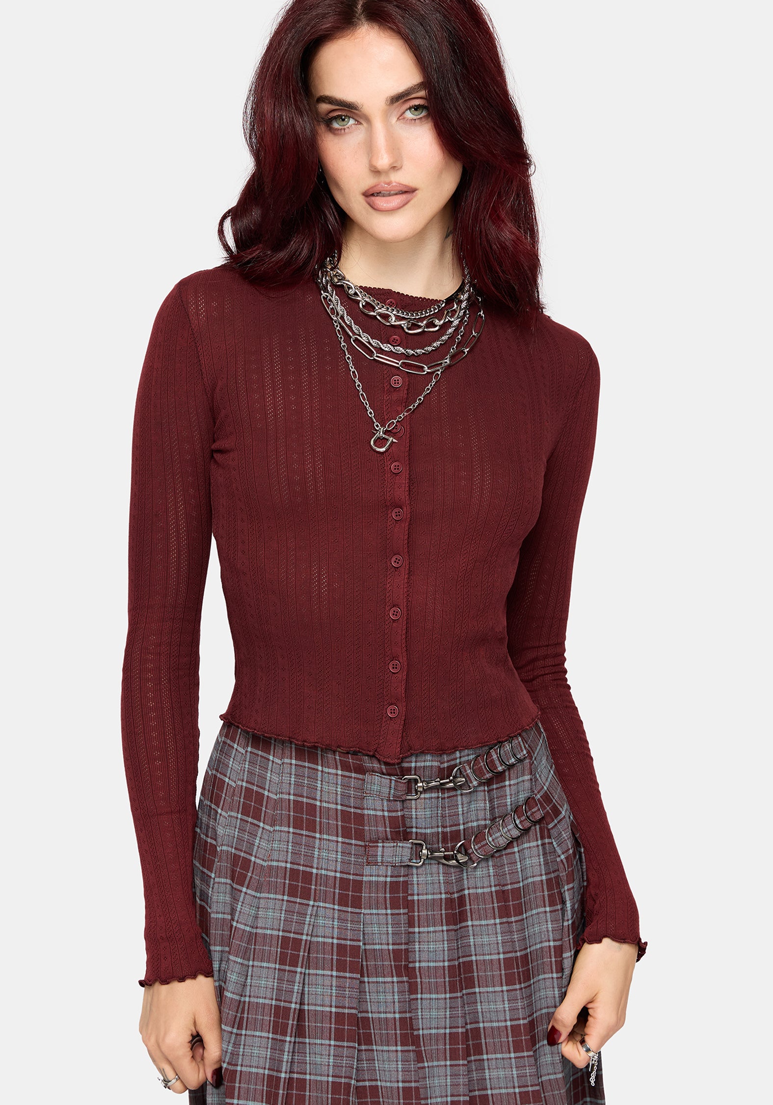A woman wearing a ribbed long-sleeve button-front knit top in deep burgundy, paired with a gray and burgundy plaid skirt and layered silver chain necklaces, against a light gray studio background.