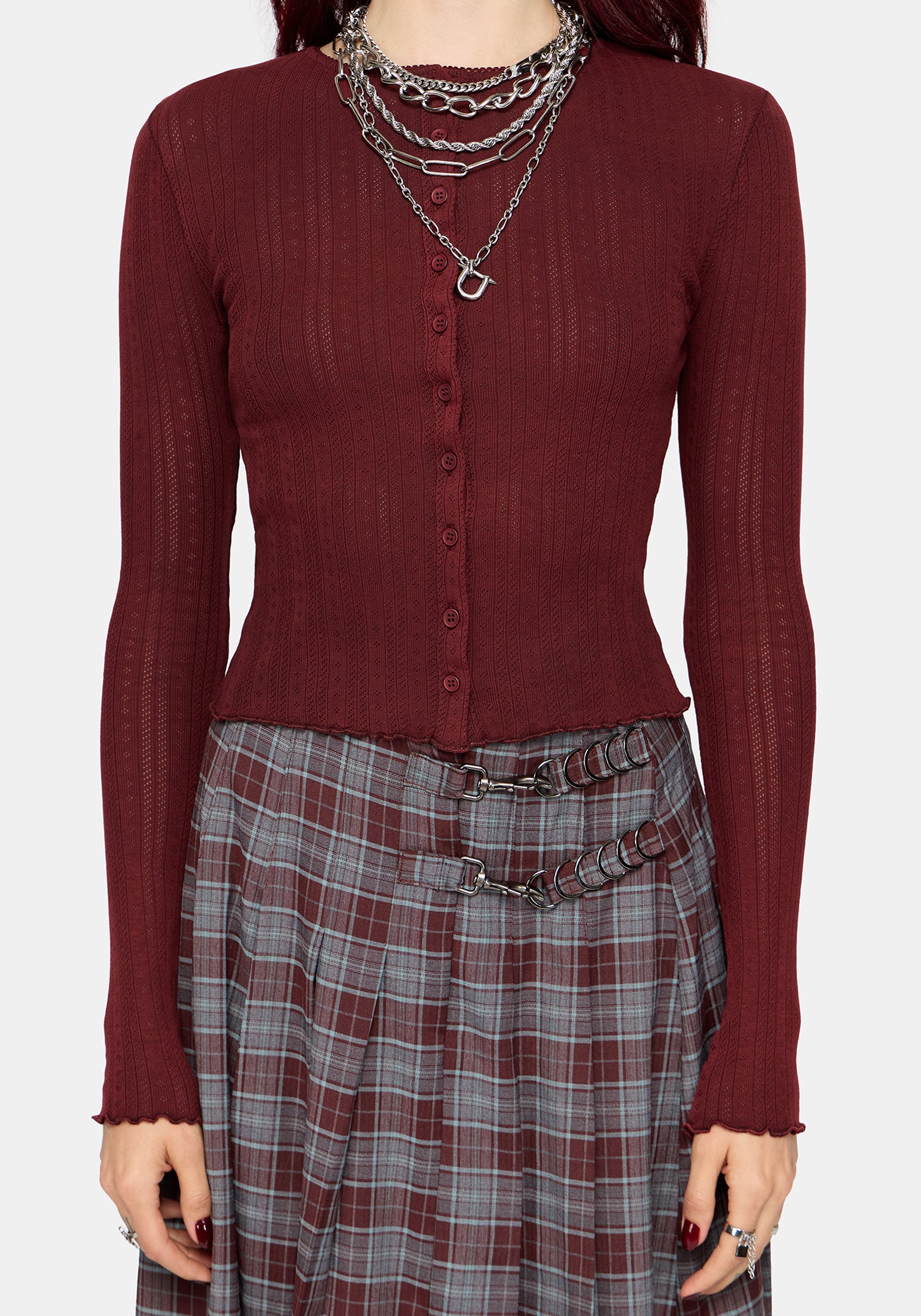 A woman wearing a ribbed long-sleeve button-front knit top in deep burgundy, paired with a gray and burgundy plaid skirt and layered silver chain necklaces, against a light gray studio background.
