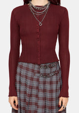 A woman wearing a ribbed long-sleeve button-front knit top in deep burgundy, paired with a gray and burgundy plaid skirt and layered silver chain necklaces, against a light gray studio background.