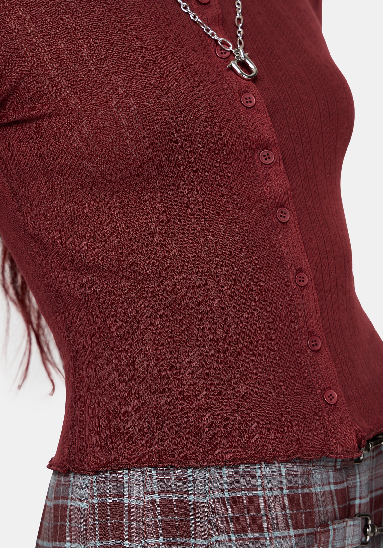 A close-up of a ribbed knit top in deep burgundy, featuring a button-down front and a silver chain necklace, paired with a plaid skirt.