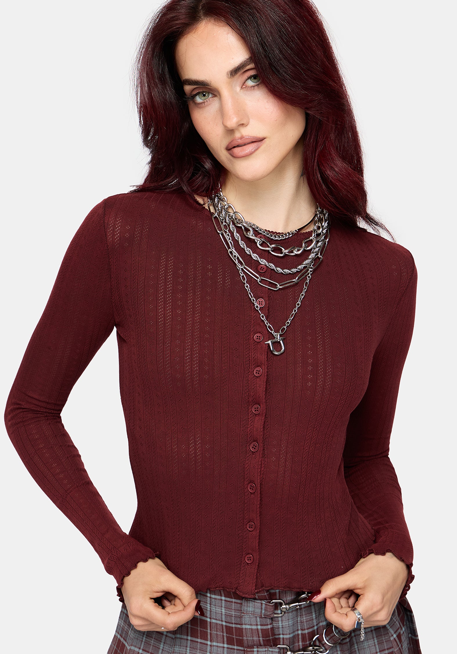 A woman wearing a long-sleeve ribbed knit button-up top in deep burgundy, styled with layered silver chain necklaces and plaid pants, against a light gray background.