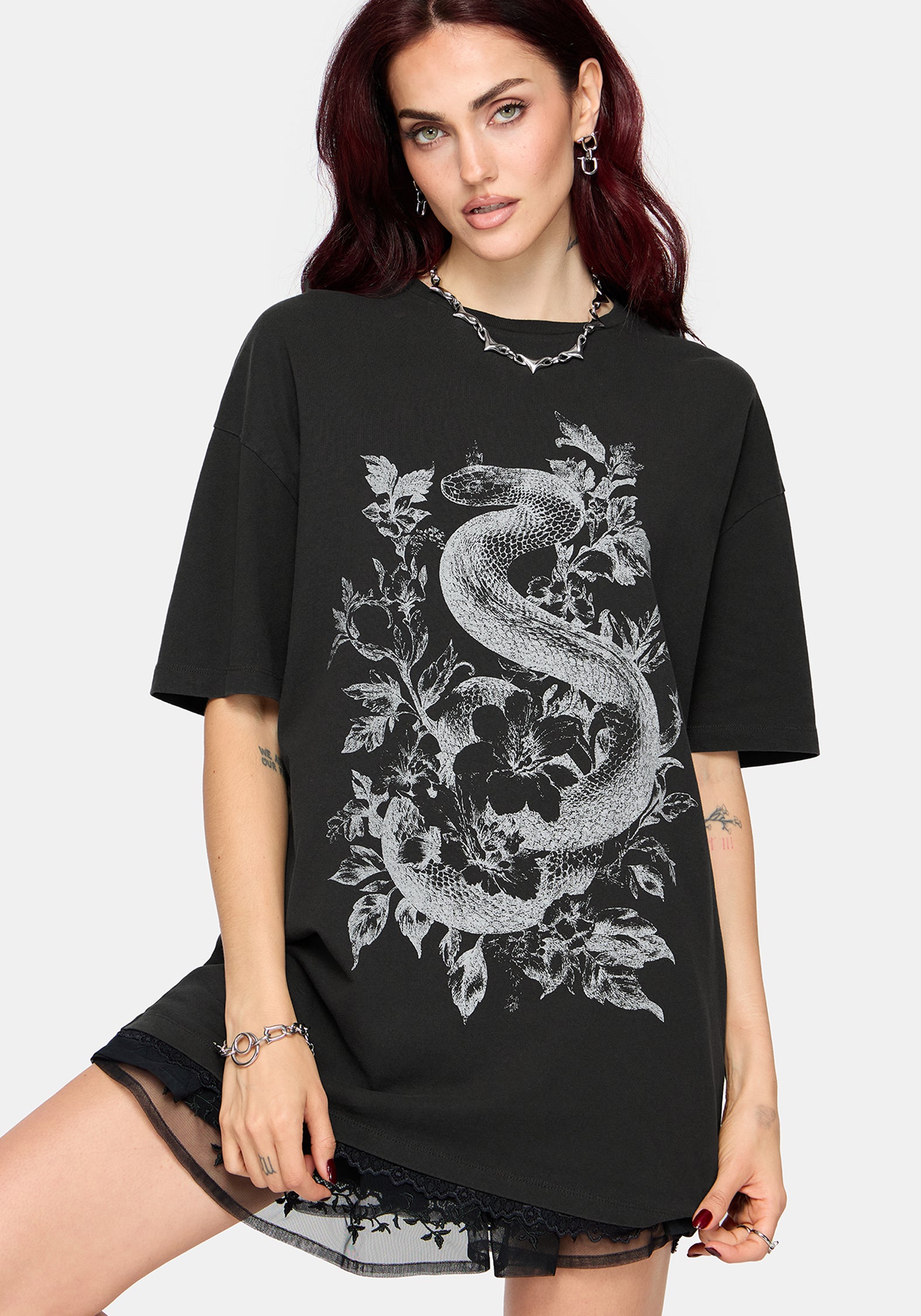 A woman modeling an oversized black t-shirt with a large white graphic print of a coiled snake and floral elements on the front, set against a plain light gray background.