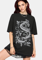 A woman modeling an oversized black t-shirt with a large white graphic print of a coiled snake and floral elements on the front, set against a plain light gray background.