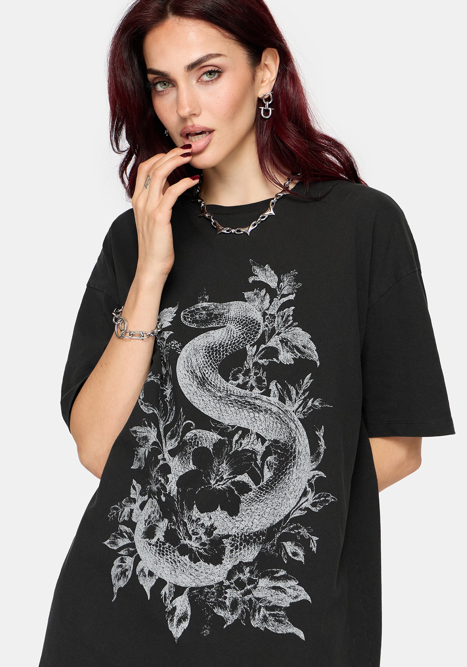 A woman wearing an oversized black t-shirt with a detailed white graphic of a snake wrapped through floral elements, set against a plain light gray background.
