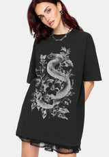 A woman wearing an oversized black t-shirt dress with a large white snake and floral graphic on the front, paired with a sheer black lace hem.
