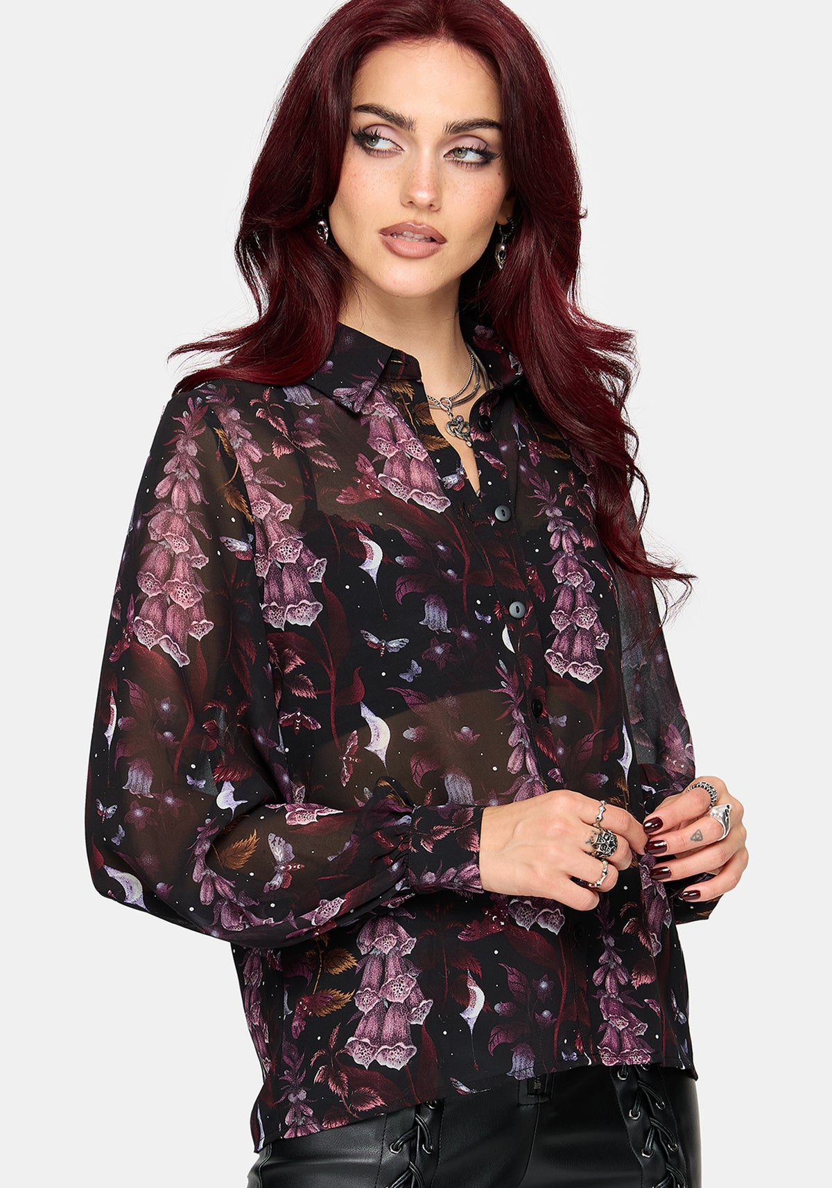 A woman with long, dark red hair models a sheer black long-sleeve blouse featuring a purple and pink floral pattern, paired with black lace-up pants, against a solid light gray background.