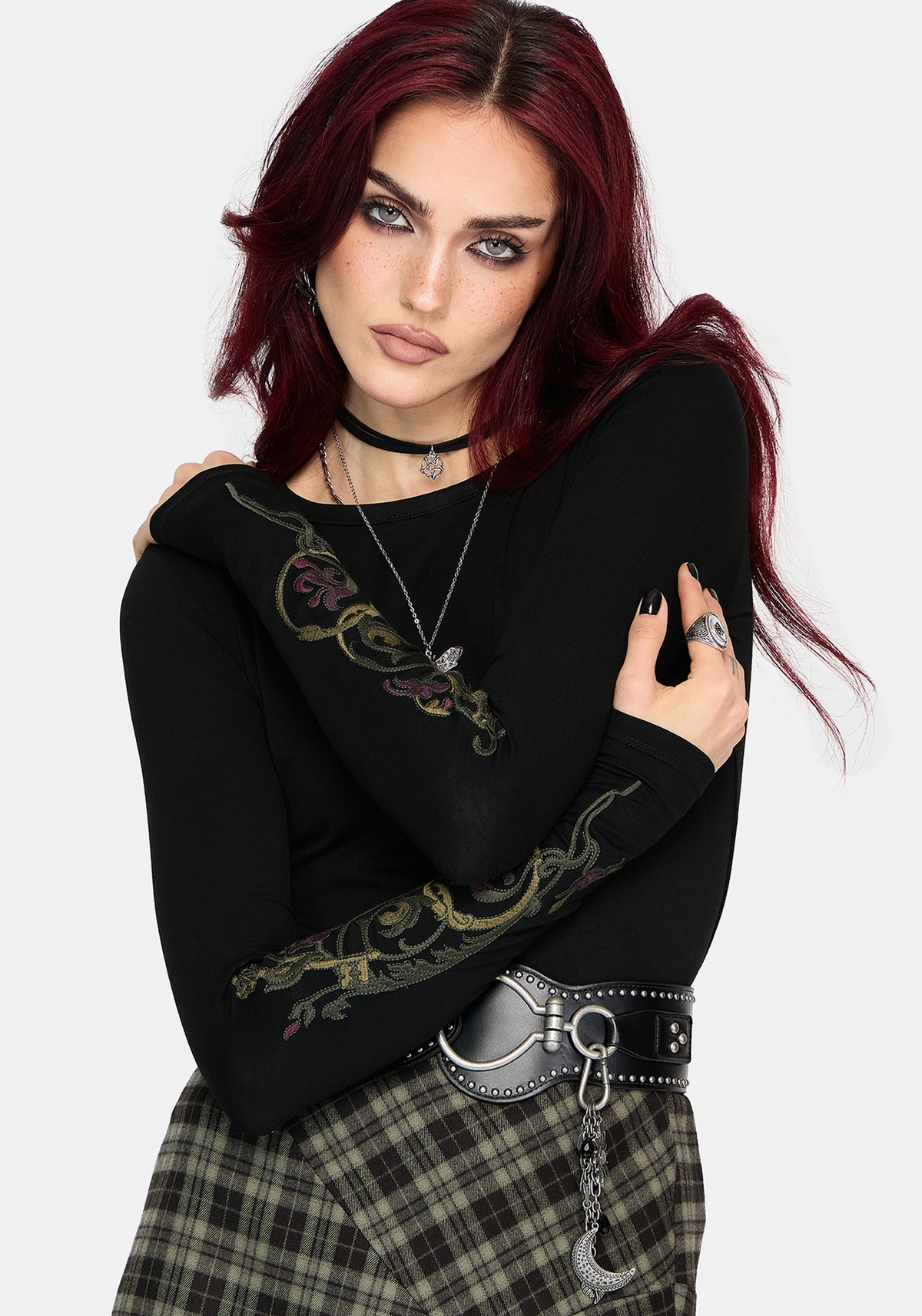 A model with dark red hair wearing a black long-sleeve top with a green and gold dragon graphic on the sleeve, paired with a plaid skirt and a silver belt.