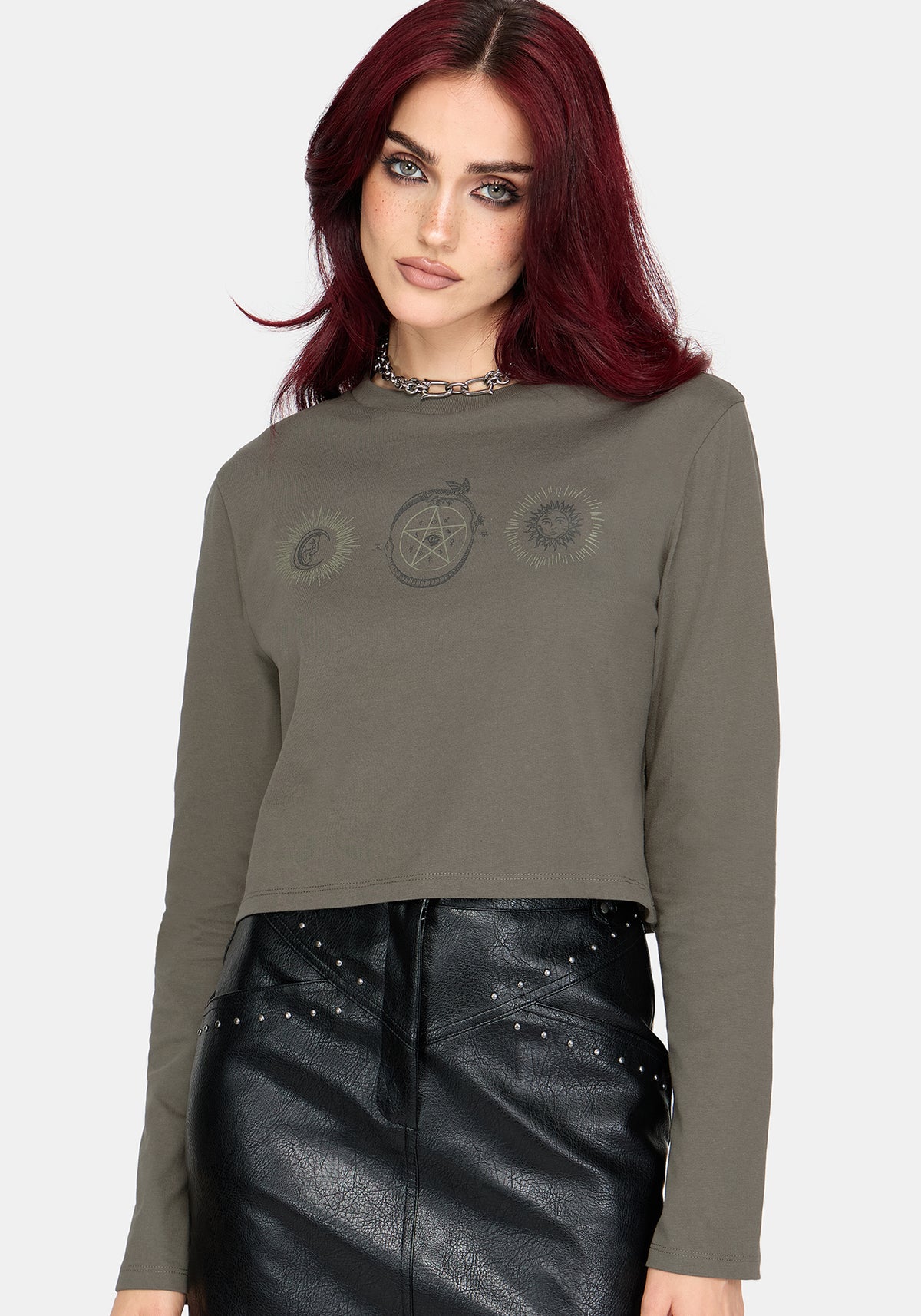 A woman with red hair wearing a cropped long-sleeve t-shirt in olive green with a gothic-style pentagram graphic on the chest, paired with a black faux-leather skirt.