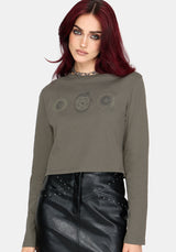 A woman with red hair wearing a cropped long-sleeve t-shirt in olive green with a gothic-style pentagram graphic on the chest, paired with a black faux-leather skirt.
