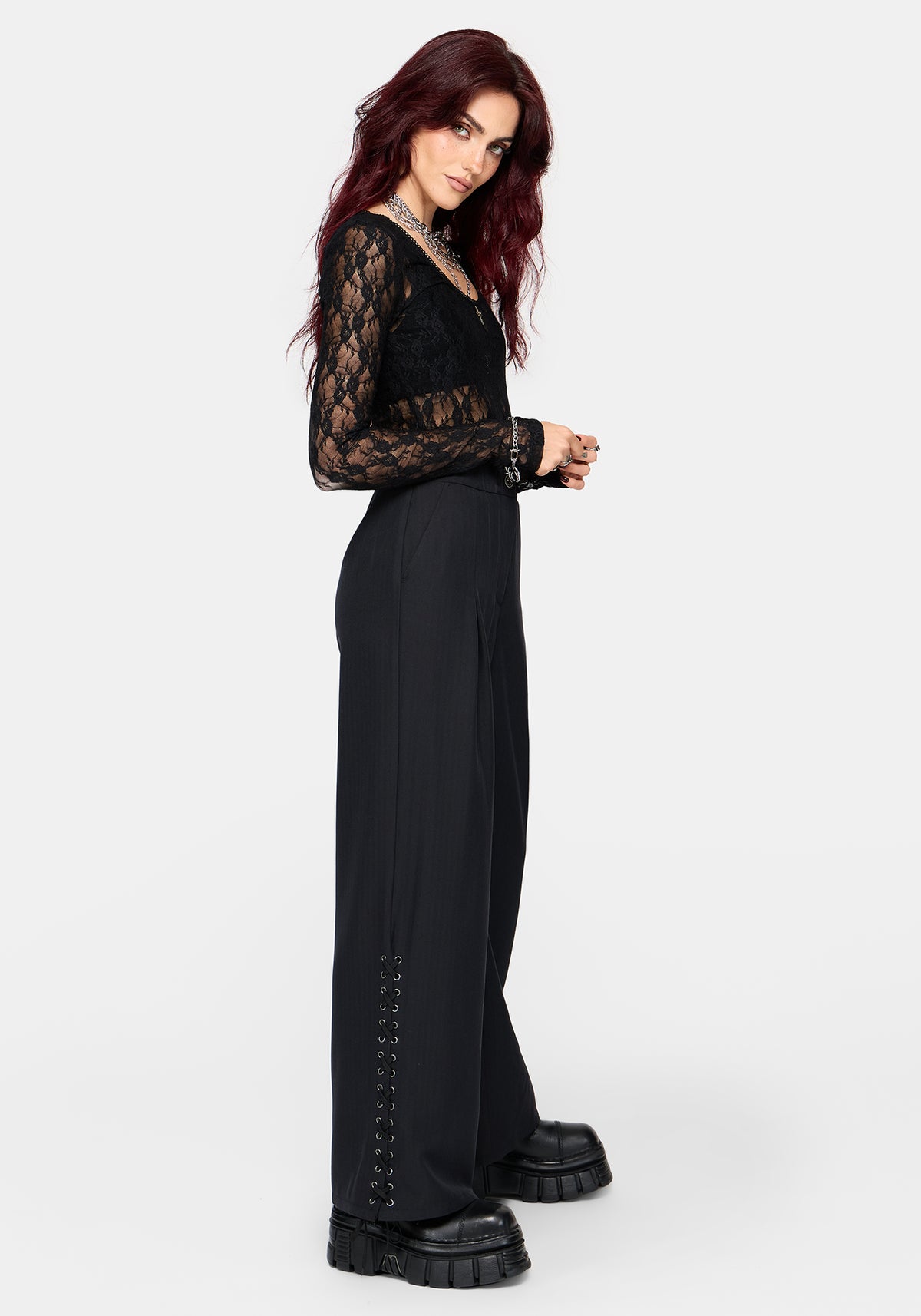 A woman modeling a black lace long-sleeve top and wide-leg black trousers with lace-up detailing down the side, paired with chunky black platform boots, against a light gray studio background.