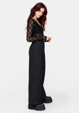 A woman modeling a black lace long-sleeve top and wide-leg black trousers with lace-up detailing down the side, paired with chunky black platform boots, against a light gray studio background.