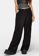 A model wearing black wide-leg pleated trousers with a button closure at the waist, styled with a gray textured crop top and chunky black platform shoes against a light gray studio background.