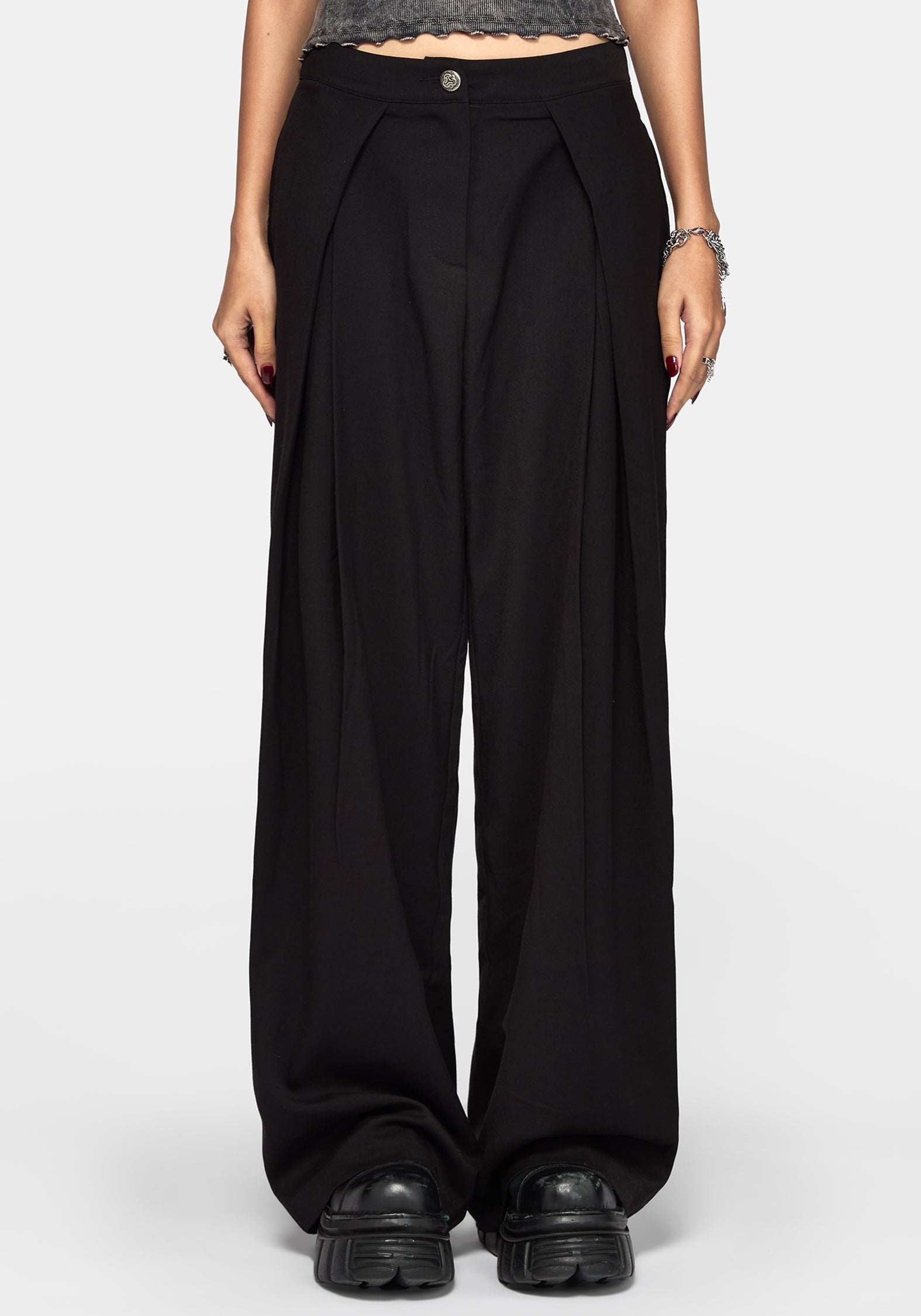 A model wearing black wide-leg pleated trousers with a button closure at the waist, styled with black platform shoes and a gray textured crop top against a light gray background.