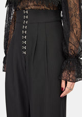 Close-up of a model wearing black high-waisted pleated trousers with functional hook and eye down the front waistband, paired with a sheer black lace top.
